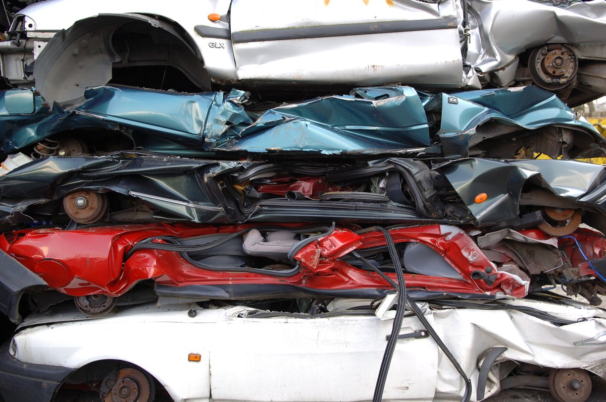 Carcasse d'auto schiacciate e impilate, carrozzerie argento, verde, nera, rossa e bianca, mozzi arrugginiti e scritta GLX